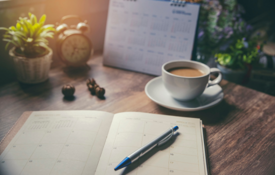 A study desk with a clock, planner, cup of coffee and plants on it. 