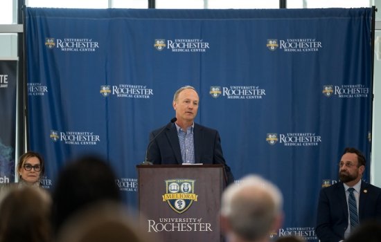 A man in a jacket stands a brown University of Rochester lectern facing an audience. 