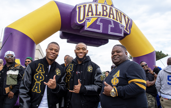 Three men in black and gold Alpha Phi Alpha fraternity jackets smile and flash shaka signs in front of a large inflated purple and gold UAlbany arch. 