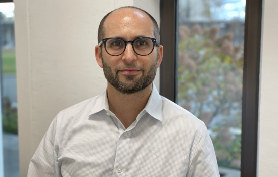 A man with close-shaved hair and glasses wears a white button-down shirt and poses for a portrait against a window.