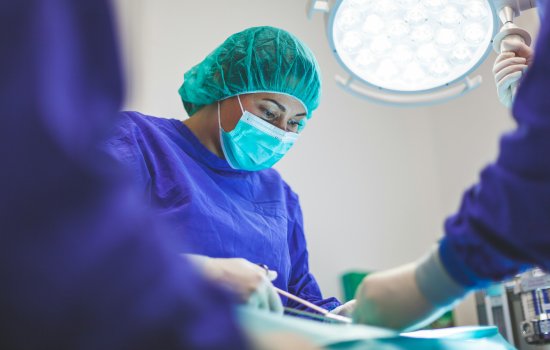 A surgeon wearing blue scrubs and a blue face mask works on a patient in a brightly lit room. 