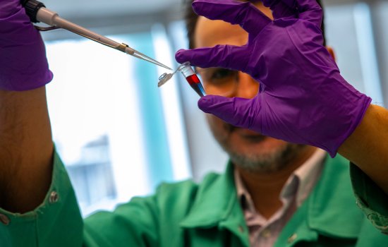 A man with brown hair and glasses holds a small plastic vial containing liquid that is purple at the bottom and red on top. He is wearing purple gloves and a green lab coat.