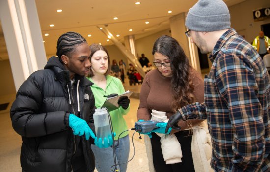 Four young people, two men and two women, stand together holding instruments to collect and record water quality measurements. One of the women is holding a gray electronic device connected to wires with probes placed in a water bottle held by a man wearing blue gloves. The second woman looks on, holding a clipboard and pen. The second man, wearing a plaid shirt instructs the group.