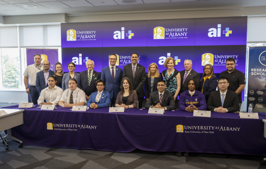 Chancellor King, along with SUNY leaders and elected officials, stand behind a group of eight UAlbany students sitting at a table branded with the UAlbany Minerva logo.