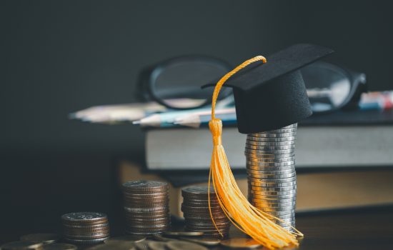 Coins stacked on a desk with books behind it, with the tallest stack of coins with a tiny graduation cap on it.