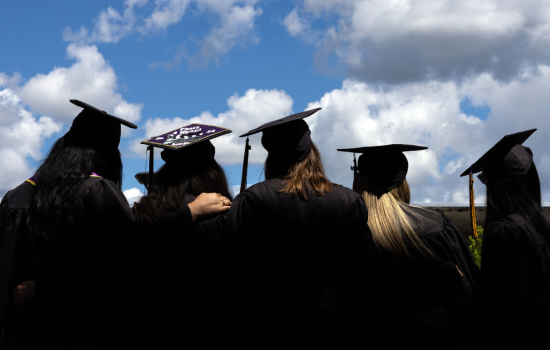 The view of the back of four UAlbany graduates stand in their caps and gowns looking at the sky.
