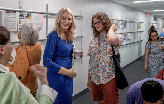 A woman with long blonde hair in a blue dress smiles in a hallway full of people who are viewing artwork on the walls.