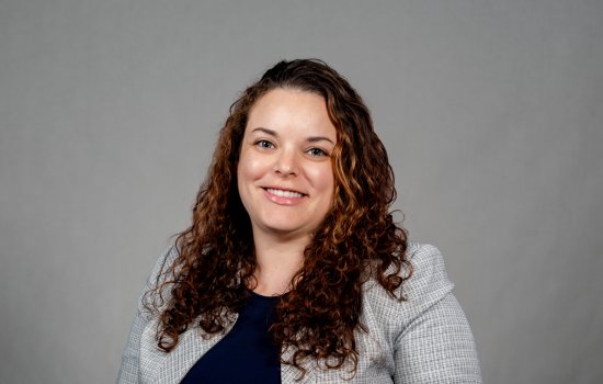 Sarah Domoff poses for a portrait against a gray background wearing a light gray jacket over a navy blue shirt. Domoff is smiling and has brown curly hair. 