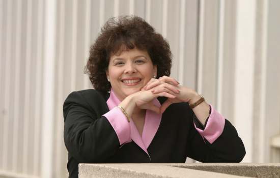 A woman with short curly hair in a pink and black blazer clasps her hands together and rests her elbows on a concrete ledge, smiling for a portrait.