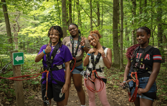 Four students wear harness gear for a high ropes course and pose for a picture in the woods.