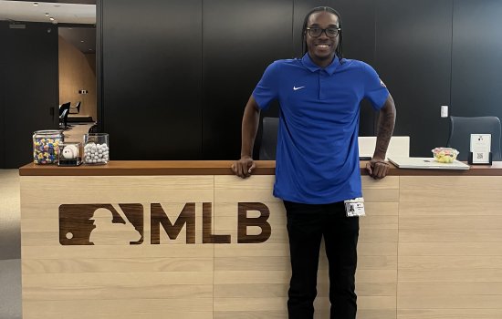 Cameron Felix stands in front of a welcome desk at MLB headquarters wearing a blue polo with his name badge attached.