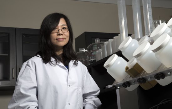UAlbany Environmental and Sustainable Engineering Professor and Chair Yanna Liang stands in her lab in a white lab coat with many containers in the foreground. 