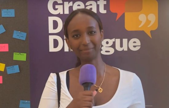 a young woman holding a microphone stands in front to a sign that reads Great Dane Dialogue