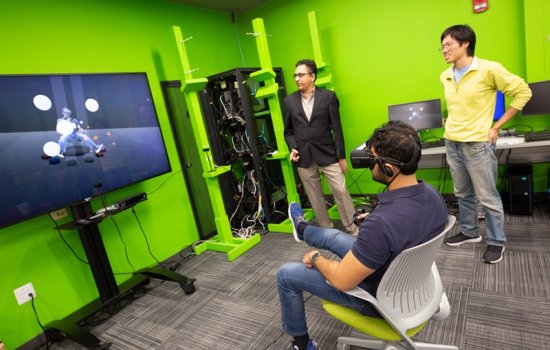 A man sitting in a chair wearing VR goggles faces a TV screen while two man stand in the background.