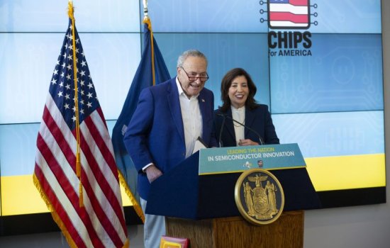 A man in a blue sport jacket and a woman in a dark business suit stand at a lectern next to an American flag. The sign on the lectern says: "Leading the Nation in Semiconductor Innovation"