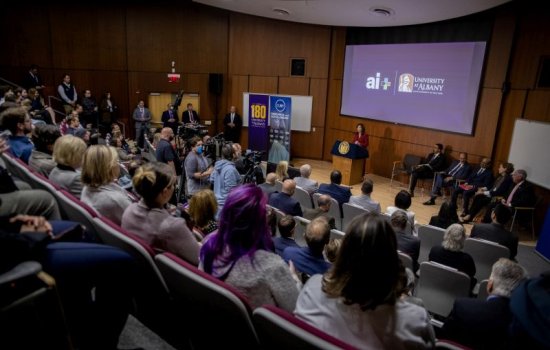 A woman in a red suit stands at a lectern in front of a packed auditorium.