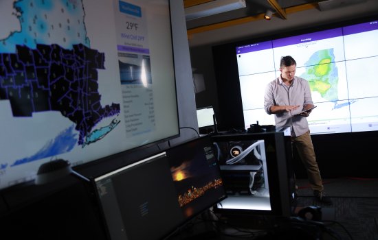 A man with an ipad looks down while a weather map of NEw York state appears in the foreground at UAlbany's XCite Lab.