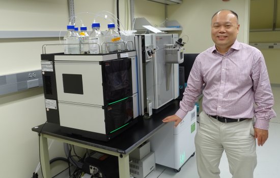 A man wearing khaki pants and a plaid button down shirt stands, smiling, in a brightly lit lab next to a DNA/RNA sequencing machine. The machine consists of two large metal cube-shaped containers with bottled of clear liquid on top of one, and a digital panel on the other. 