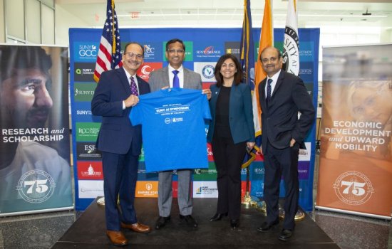 Three men and a woman stand shoulder-to-shoulder in front of a blue SUNY backdrop smiling and holding a blue t-shirt that reads "SUNY AI Symposium"