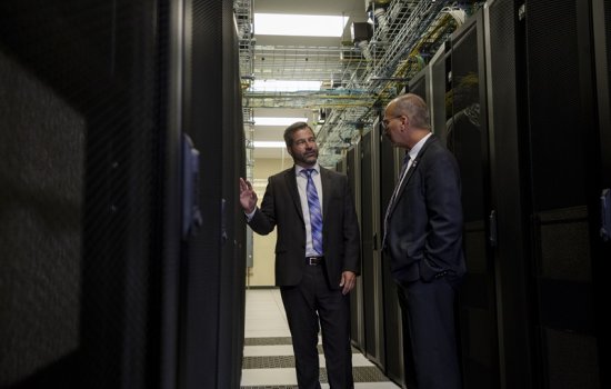 Two men in dark suits stand facing one another between two long rows of computer racks.