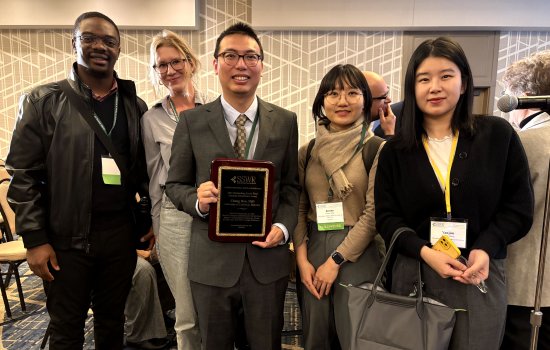 Five people - two men and three women - pose together, smiling, for a portrait. They are standing in a room with geometric-printed wallpaper and carpeting. All are wearing badges on lanyards to signify participation in the Society for Social Work and Research annual meeting.