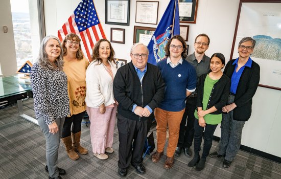Eight people, including five women and three men smile together for a group portrait in an office with gray carpet and a collection of framed plaques in the background. An American flag and the flag of New York State stand behind the group.