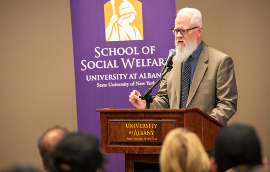 Eric Hardiman behind a podium. Behind him is a purple banner that says "School of Social Welfare".