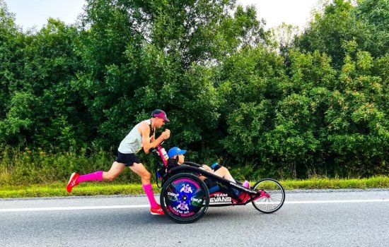 A runner pushes a peron seated in a racing chair along a road with greenery in the background