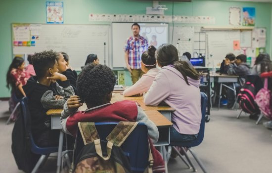 Elementary school students sit in a classroom and stare at a man in the front of the class.