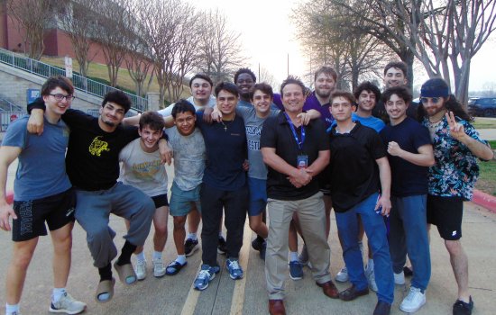 Members of the UAlbany wrestling team, including 15 young men, pose for a team photo outside of an arena in a roadway.