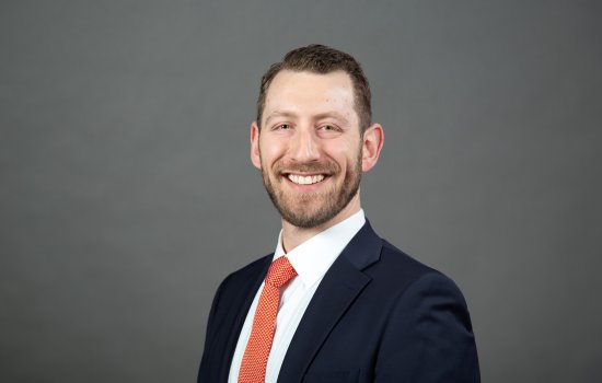 A man wearing a dark blue suit and dotted red tie with close-cropped brown hair and a full beard smiles at the camera in front of a gray background.