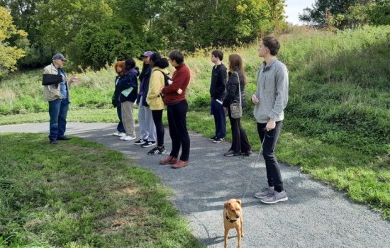 A group of students standing along a nature path listen as a man in a sling and ballcap talks. One students holds a dog on a leash.