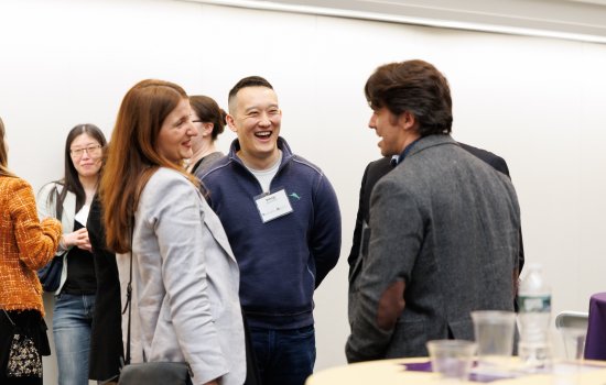 A group of people stand and talk at an indoor event; some are smiling and laughing, with name tags visible on a few attendees.