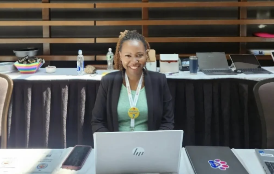 . A woman at a table uses her laptop, with various papers organized around her.