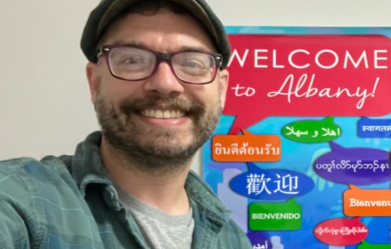 Daniel Butterworth poses for a selfie in front of a sign that reads, "Welcome to Albany!" in English and several other languages.