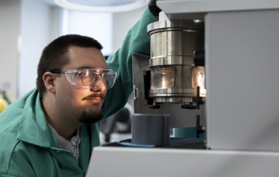 A man wearing glasses, protective goggles and a green lab coat handles a lab instrument that features a metal cylinder containing a lit glass chamber. 
