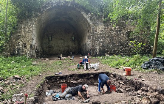 Archaeologists work in a hole digging with tools, with one person recording notes, in the background is an archway of a ruined church from the 1500s.
