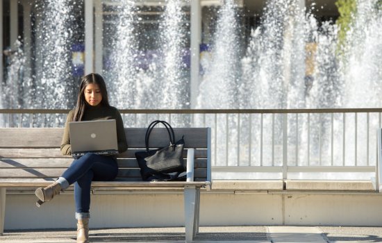 Student on laptop sitting on a bench in front of fountain