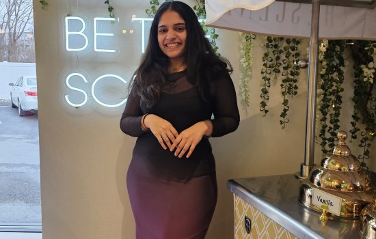 Smiling woman in a black and purple dress stands beside a vintage coffee cart. Neon sign and green vines hang on the wall, creating a cozy ambiance.