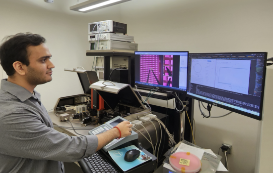 A man with short dark hair in a gray shirt stands in front of two computer screens in a laboratory.