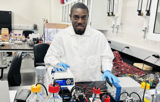 A man with short black hair and a beard wears a white lab coat and blue glove and touches equipment in a laboratory.