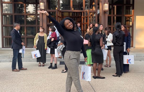 Alissa Banfield poses with a bag in one hand and an arm in the air outside of a building where a crowd has gathered.