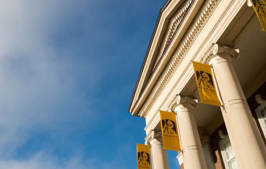 A bright blue sky accentuates the roof of a brick building with ornate columns holding yellow UAlbany flags