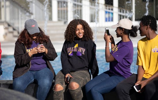 A student smiles and poses for photo while sitting on the edge of UAlbany's fountain with three friends. All four students are wearing UAlbany t-shirts or sweatshirts.