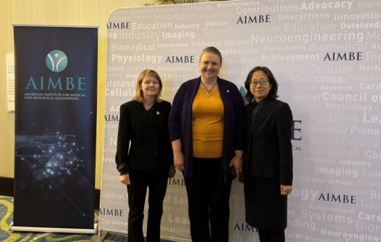 Three women smile for a photo, standing against a backdrop printed with the name “AIMBE” in purple, with a collection of science-related words in very faint print. To their left is a pull-up poster that says “AIMBE American Institute for Medical and Biological Engineering."