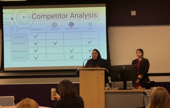 Two students stand at the front of a college classroom and present a slide with the title Competitor Analysis.