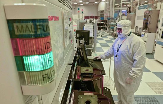 A man works at a computer terminal inside of a high tech clean room wearing a full white cleanroom suit, including white gloves and a white cap.