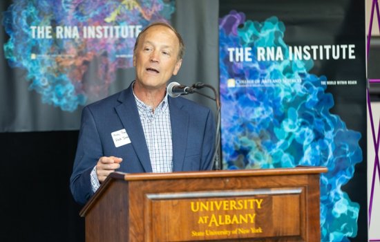 A man speaking at brown wood lectern with the words "University at Albany" written on the front in gold, in front of a colorful banner that says "The RNA Institute."