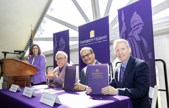 Four people are pictured under a white event tent with strong sunshine coming through. A woman wearing a purple blazer stands at a wooden podium. Three men wearing blazers sit at a table with a purple tablecloth, all smiling at the camera. Purple “University at Albany” banners are behind them.