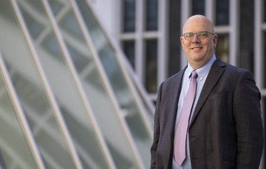 A smiling man in a gray jacket and purple tie stands outdoors near windows